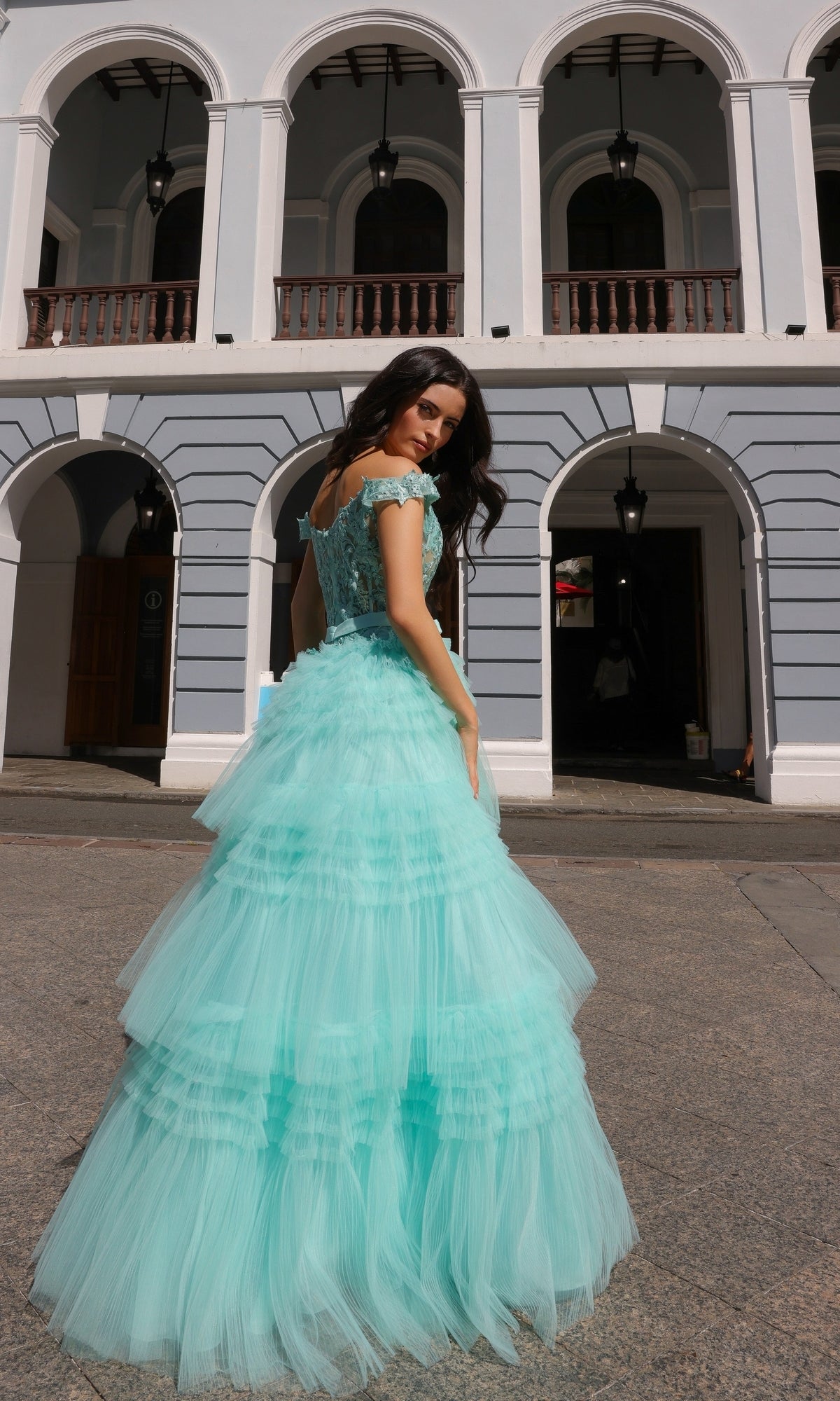 A woman poses outdoors in front of a gray and white building wearing the Nox Anabel Designer Ruffled Prom Ball Gown E1293, a light blue layered tulle dress.